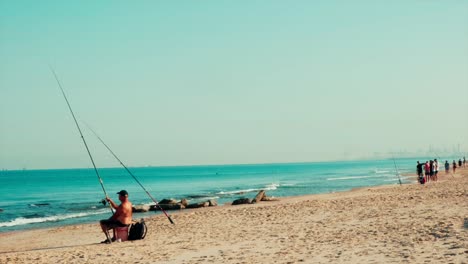 Statische-Aufnahme-Eines-Alten-Mannes-Ohne-Hemd-Beim-Angeln-Am-Strand,-Menschen-Im-Hintergrund