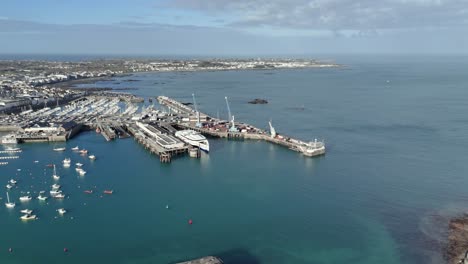 Vuelo-De-St-Peter-Port-Guernsey-Sobre-La-Terminal-De-Ferry-Con-Ferry-En-El-Muelle,-Muelle-Comercial-Con-Grúas-Con-Qe-Ii-Marina-En-El-Fondo-Y-Vistas-A-La-Bahía-De-Belle-Greve-En-Un-Día-Soleado