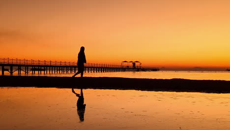 Silhouette-Einer-Frau,-Die-Am-Strand-Spazieren-Geht,-Mit-Gespiegelter-Reflexion-Auf-Dem-Wasser,-Im-Hintergrund-Ein-Holzsteg-Bei-Sonnenuntergang-Zur-Goldenen-Stunde