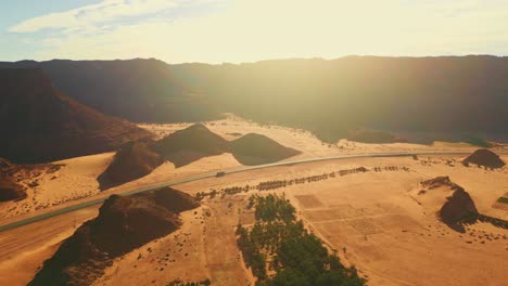 Carretera-Con-Tráfico-Ligero-En-Carretera-Asfaltada-A-Través-Del-Desierto-Con-Palmeras-Verdes,-Cañón-Con-Montaña-De-Roca-Naranja-Y-Arena-Amarilla-Al-Atardecer