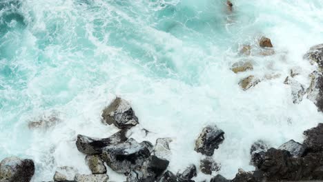 water-waves-crashing-towards-the-large-rocks-on-the-seashore,-static-top-view