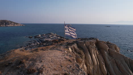 National-Flag-of-Greece-Waving-on-Pole-on-Top-of-Cliff-Above-Aegean-Sea,-Drone-Shot