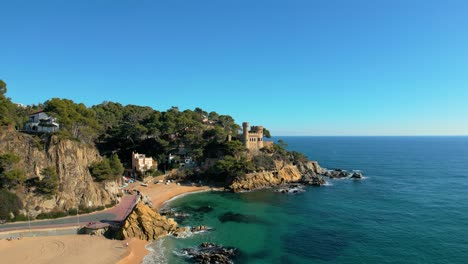 Vistas-Aéreas-De-La-Playa-Principal-De-Lloret-De-Mar,-Castillo-Al-Fondo,-Amplia-Playa-Sin-Gente,-Arena-Gruesa,-Agua,-Azul-Turquesa