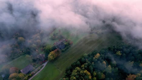 An-aerial-view-of-a-dense,-green-forest-with-patches-of-sunlight-breaking-through-the-fog,-revealing-several-houses-with-brown-roofs-nestled-among-the-trees