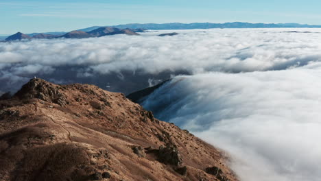 Pico-De-Montaña-Que-Se-Eleva-Sobre-El-Mar-De-Nubes-Bajo-Un-Cielo-Azul-Claro,-Figura-Solitaria-En-La-Distancia