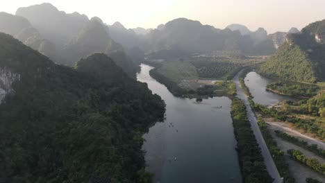 Drone-aerial-view-in-Vietnam-flying-over-a-valley-surrounded-by-rocky-mountains-covered-with-green-trees-over-a-river-in-Ninh-Binh-on-a-sunny-day