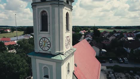 A-clock-tower-with-red-roof-in-a-quaint-village-on-a-sunny-day,-green-landscape-in-the-background,-aerial-view