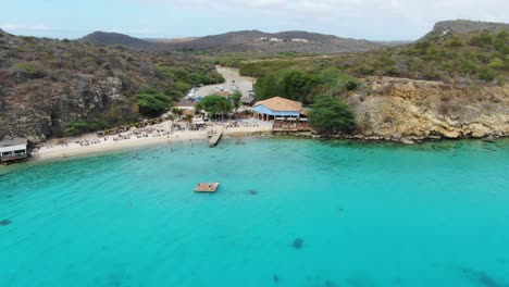 Kokomo-beach-in-curacao,-clear-turquoise-waters-with-beachgoers-and-lush-hills,-aerial-view