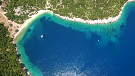 Crystal-blue-waters-edge-a-lush-green-cove-on-Kefalonia-Island,-aerial-view,-sunny-day