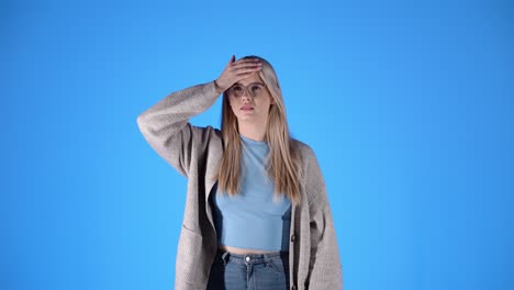 Sick-woman-checks-forehead-for-fever-and-sneezes-into-tissue,-blue-background