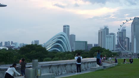 Parque-Urbano-En-La-Azotea-De-La-Presa-Del-Puerto-Deportivo-Y-Presa-De-Agua,-Toma-Estática-Que-Captura-A-La-Gente-Haciendo-Un-Picnic-Y-Pasando-El-Rato-En-La-Hierba-Verde-Con-La-Rueda-De-Observación-Del-Volante-De-Singapur-Y-Los-Jardines-Junto-A-La-Bahía-Al-Fondo