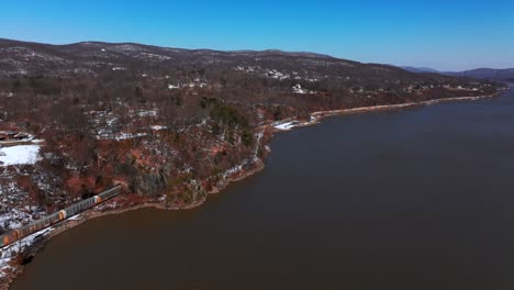 Eine-Luftaufnahme-Eines-Langen-Güterzuges,-Der-An-Einem-Sonnigen-Tag-Mit-Klarem-Blauen-Himmel-Den-Hudson-River-Entlang-Fährt