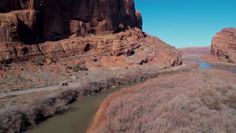 A-tracking-drone-shot-of-a-truck-driving-between-the-steep-cliffs,-known-as-the-“Wall-Street-Climbing-area”,-and-the-Colorado-River,-near-Moab,-Utah