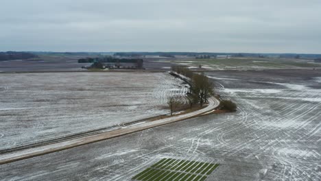 Fly-above-countryside-gravel-road-with-trees-and-frozen-agriculture-grain-field