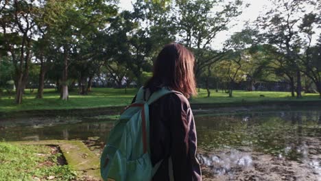 Joven-Asiática-Con-Mochila-Caminando-Junto-Al-Estanque-En-El-Parque