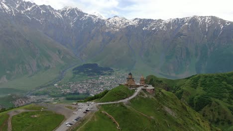 Drone-aerial-view-in-Georgia-flying-sideways-Gergeti-Trinity-orthodox-church-in-Kazbegi-surrounded-by-green-mountains-valley-with-snowed-peaks