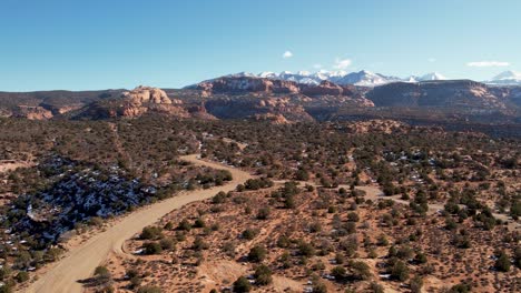 Un-Dron-De-Alto-Vuelo-Disparado-Sobre-Un-Remoto-Camino-De-Tierra-Que-Atraviesa-La-Vasta-Y-única-Tierra-Desértica-Cerca-De-Moab,-Utah,-Con-Las-Montañas-Rocosas-Nevadas-Que-Se-Elevan-En-La-Distancia