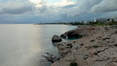 Bridge-of-Love-or-Love-Bridge-in-Cyprus-At-Twilight-with-Dramatic-Sky-at-Cape-Greco,-Ayia-Napa-Town-Coastline-in-Background---Aerial-pull-back-towards-cliff-edge