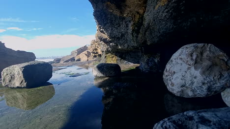 Pequeñas-Gotas-Que-Caen-De-Las-Cuevas-Formadas-Por-El-Mar-En-La-Ladera,-Forman-Un-Pequeño-Espejo-De-Agua-Contra-El-Cielo-Azul