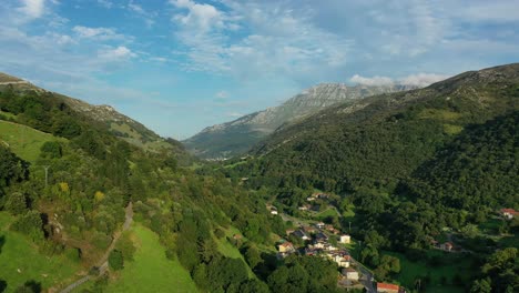 Vuelo-En-Un-Valle-Descendiendo-En-Un-Pueblo-Con-Sus-Casas-Rurales-Laderas-De-Verdes-Prados-Y-Bosques-Con-Un-Fondo-De-Una-Montaña-De-Rocas-Calizas-Con-Un-Cielo-Azul-Con-Nubes-Cantabria-españa