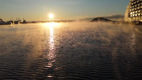 Floating-Fog-Moving-Over-Water-Beside-Apartments-With-Golden-Hour-Sunset-In-Background-In-Bjorvika,-Oslo