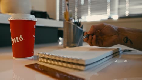 A-low-angle-shot-of-an-artist's-hand-sketching-on-a-notepad,-featuring-a-foreground-coffee-cup