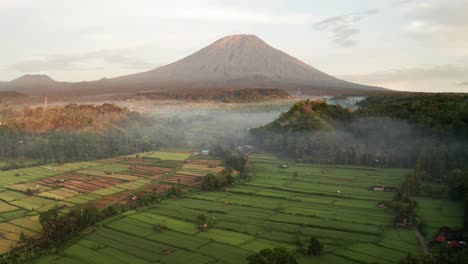 Toma-Inversa-De-Drones-Del-Volcán-Monte-Agung-Con-Niebla-Matutina-Y-Exuberantes-Campos-De-Arroz-En-Bali,-Indonesia