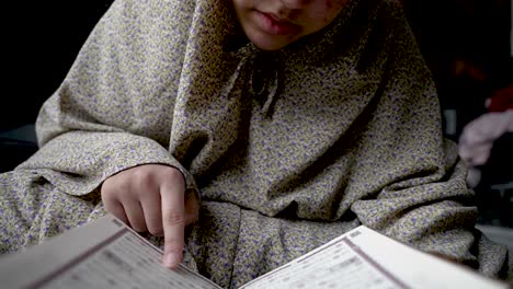 Child-in-traditional-attire-engrossed-in-reading-the-Quran,-indoors,-intimate-close-up