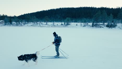 Ein-Mann-Fährt-Mit-Seinem-Alaskan-Malamute-Ski-In-Einer-Verschneiten-Landschaft---Kamerafahrt