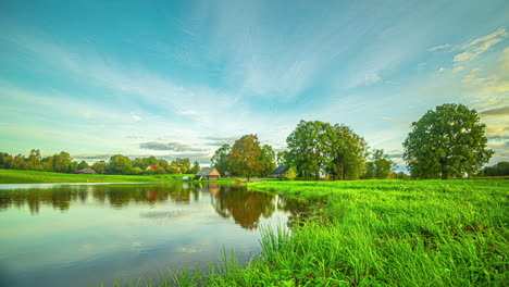 Goldene-Wolken-Ziehen-über-Den-Blauen-Himmel-Und-Spiegeln-Sich-Im-Ruhigen-Teich,-Während-Der-Wind-Gras-Und-Bäume-In-Bewegung-Versetzt