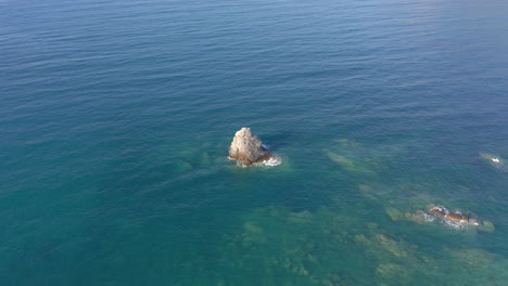 Aerial:-Slow-panning-drone-shot-of-a-big-rock-in-the-middle-of-the-blue-clear-and-tranquil-sea