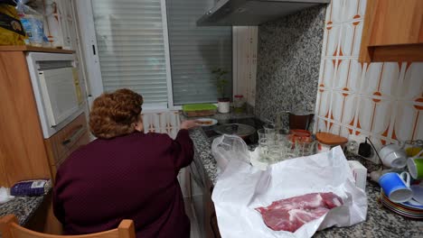 Elderly-lady-preparing-to-cook-meat-in-her-kitchen-seated-on-a-chair