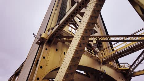 Low-angle-footage-of-crossing-beams-on-a-rusty-yellow-bridge-in-downtown-sacramento,-california-with-a-grey-sky-in-the-background