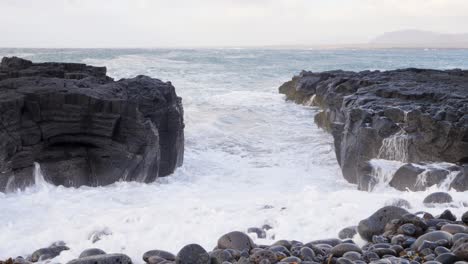 Waves-crash-against-volcanic-basalt-columns-on-an-Icelandic-beach,-pebbles-in-foreground