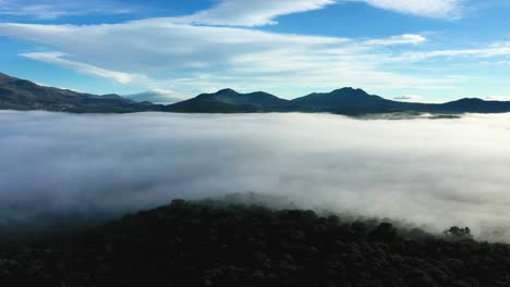 Flug-In-Ein-Meer-Von-Wolken-Im-Morgengrauen-Mit-Einem-Spektakulären-Hintergrund-Von-Bergen-Mit-Einer-Dunklen-Farbe-Aufgrund-Des-Lichts-Und-Einem-Blauen-Himmel,-Der-Einen-Bewaldeten-Hügel-Mit-Wolken-Im-Winter-Spaniens-Hinterlässt