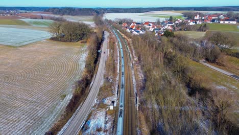Aerial-Shot-of-Train-Gliding-Past-Village-Amidst-Winter's-Fading-Snow