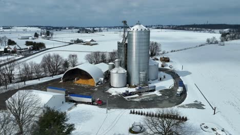 Aerial-approaching-shot-of-industrial-silo-on-farm-with-transporting-trucks-in-winter