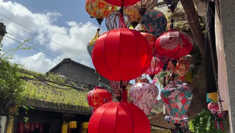 Lanterns-hanging-outside-a-shop-blowing-in-the-breeze-with-a-temple-roof-and-blue-sky-in-the-background-in-Hoi-An,-Vietnam-ancient-town