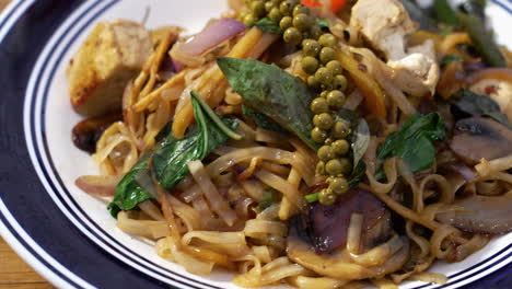 Tofu-Pad-Kee-Mao-topped-with-fresh-thai-basil-and-green-peppercorns-on-blue-and-white-plate-on-tabletop,-close-up-slider-4K