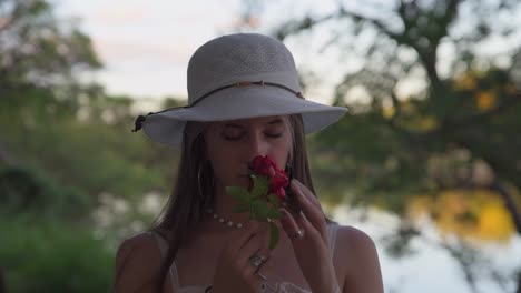 Beautiful-young-woman-with-hat-smelling-rose-while-sighing-carefree-near-lake-in-nature-at-sunset,-close-up