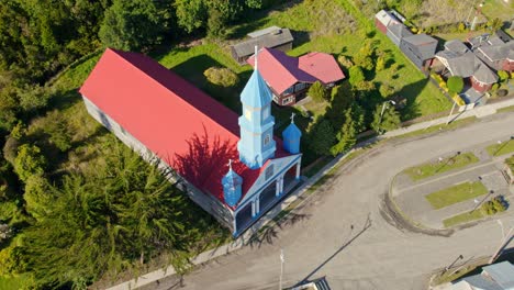 Vibrant-blue-Patrimony-church-in-Tenaun-with-a-red-roof-in-chiloé,-chile,-on-a-sunny-day,-aerial-view