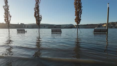 Tilting-up-for-flooded-benches-and-trees-at-the-quay-in-Budapest-with-Margaret-Bridge-in-the-background-during-the-flooding-of-Danube-River,-Hungary---December-28,-2023