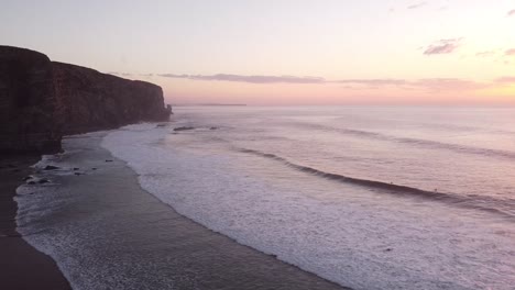drone-shot-with-the-coast-with-cliffs-at-sunset-and-surfers