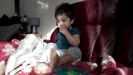 British-Asian-18-month-old-baby-boy-sits-on-a-sofa,-happily-eating-sliced-apples-in-the-morning