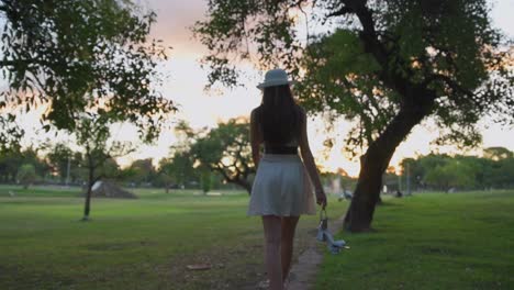 Pretty-young-woman-in-hat-walking-barefoot-carefree-through-park-carrying-shoes-and-rose-in-her-hands