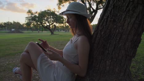 Beautiful-young-woman-with-hat-leaning-on-tree-carefree-while-looking-at-her-smartphone-in-nature-at-sunset