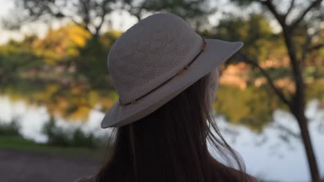 Beautiful-young-woman-walking-carefree-near-lake-while-putting-on-white-hat-in-nature-at-sunset,-close-up