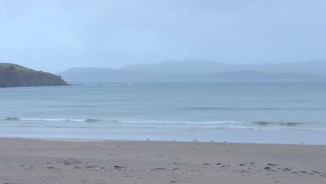 Gentle-waves-on-a-sandy-beach-with-misty-hills-in-the-distance,-serene-atmosphere,-cloudy-Irish-coastline
