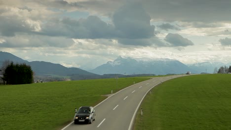 Cars-drive-on-a-highway-with-lush-green-fields-and-snow-capped-mountains-in-the-backdrop,-partly-cloudy-sky-above