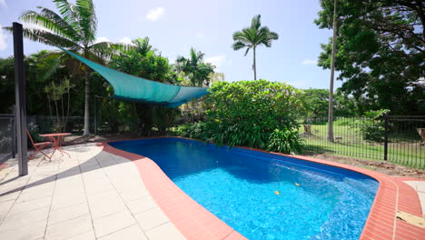 Pullback-Across-Large-Outdoor-Pool-Area-Shimmering-Reflecting-Sunlight-With-Shade-Cloth-in-Fenced-Area